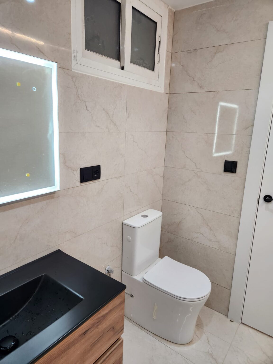 Baño moderno reformado con lavabo negro y espejo retroiluminado en Barcelona